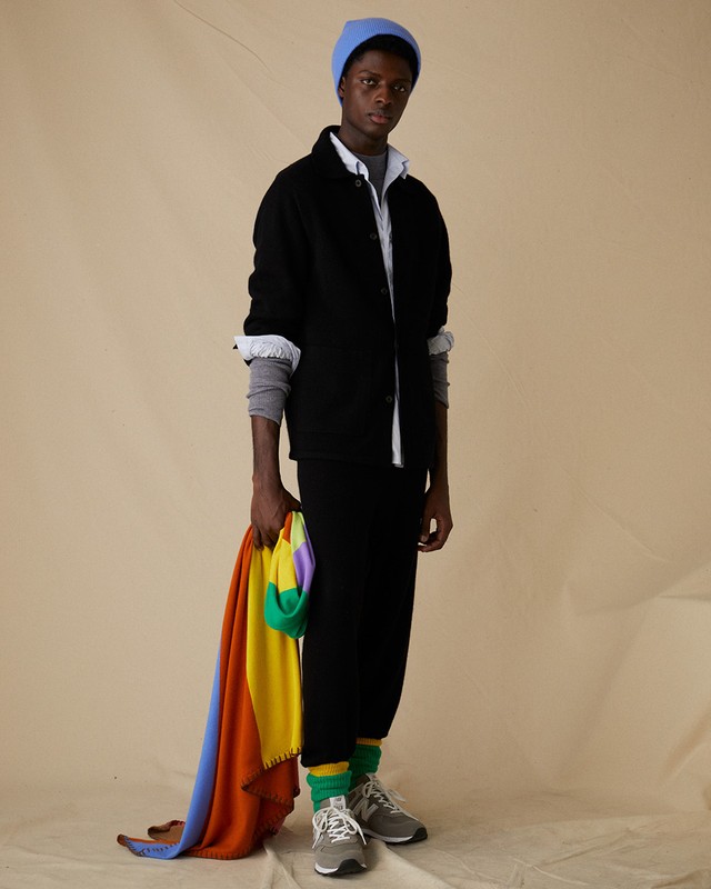 Young Black man in black casualwear and a blue beanie poses holding a colorful blanket in a studio setting with neutral background.