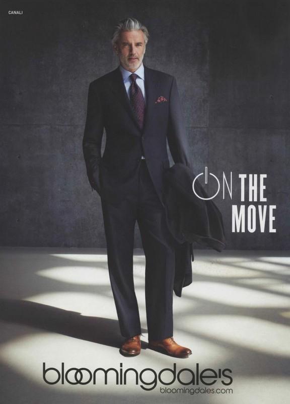 Older man in a dark suit, tie, and brown dress shoes standing against a textured grey wall with light patterns on the floor.