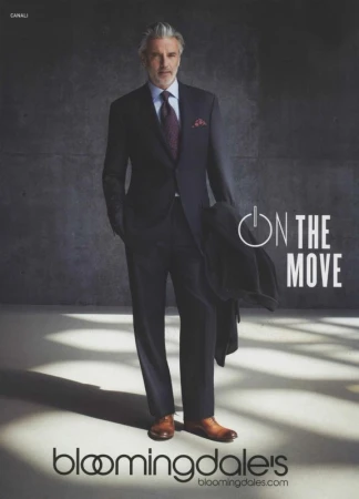 Older man in a dark suit, tie, and brown dress shoes standing against a textured grey wall with light patterns on the floor.