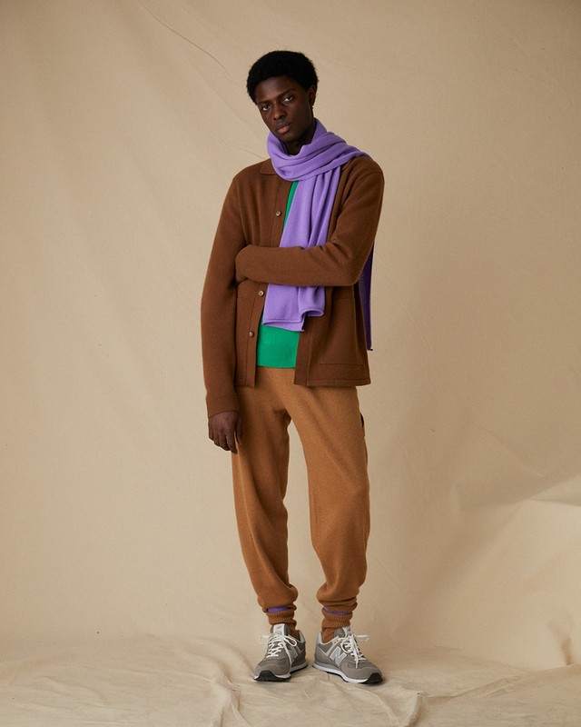 A young adult male model poses in a brown cardigan, green t-shirt, tan sweatpants, grey sneakers and a purple scarf on a beige background.