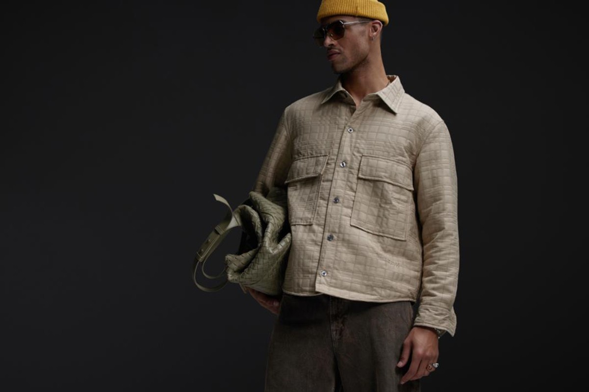 A stylish adult male in a button-down shirt, jeans, and a yellow beanie, carrying a tote bag against a dark grey studio background.
