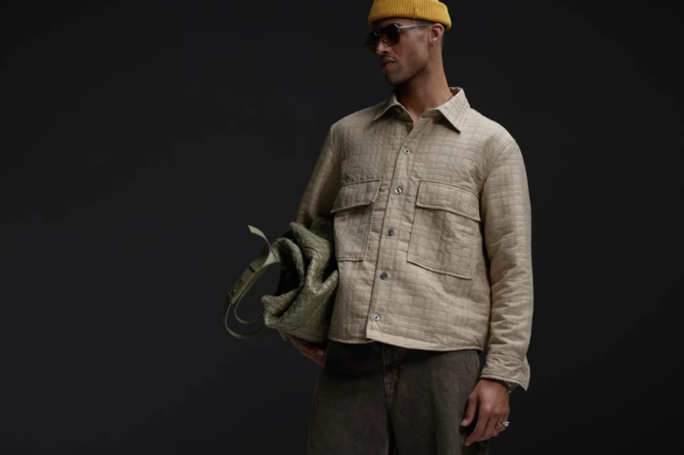 A stylish adult male in a button-down shirt, jeans, and a yellow beanie, carrying a tote bag against a dark grey studio background.