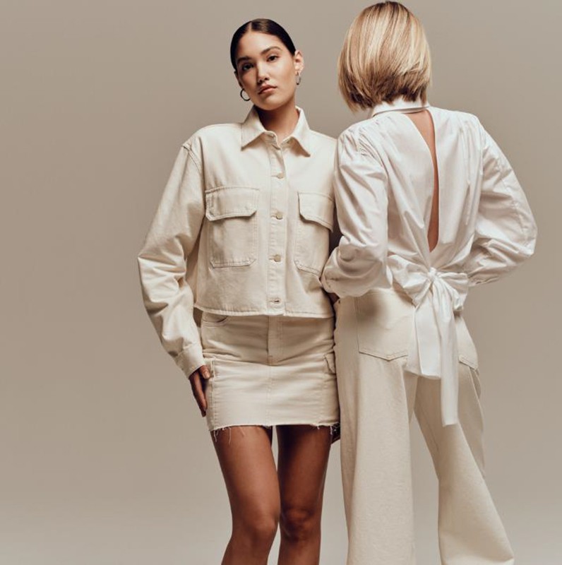 Two fashion models pose in a photo studio wearing matching white outfits: a jacket with skirt and blouse with trousers.