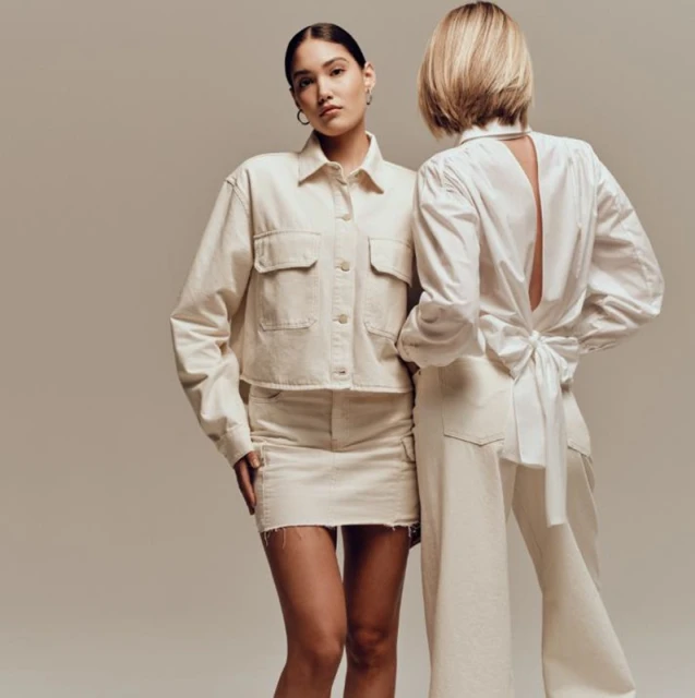 Two fashion models pose in a photo studio wearing matching white outfits: a jacket with skirt and blouse with trousers.