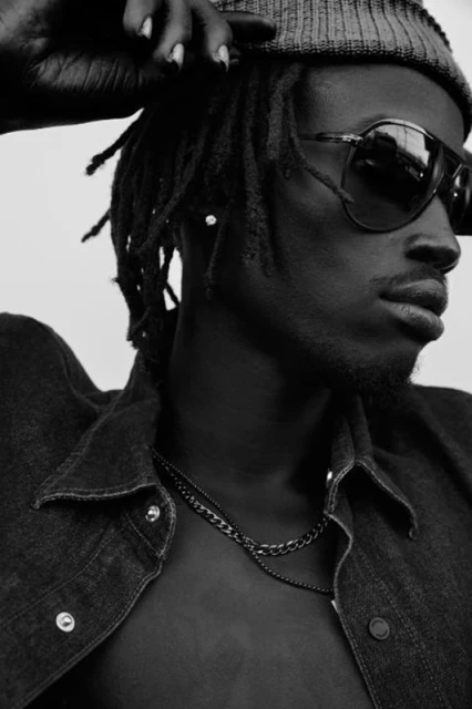 Black and white portrait of a man wearing sunglasses and a denim jacket with chain necklaces, shot in a photo studio.