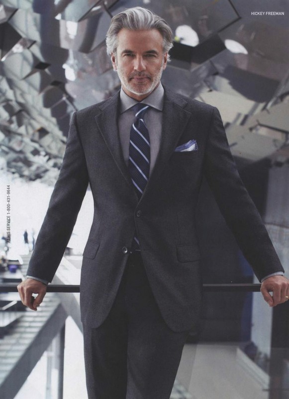 Portrait of a mature man in a dark grey suit, light grey shirt, and blue tie. He stands in an open, bright room.
