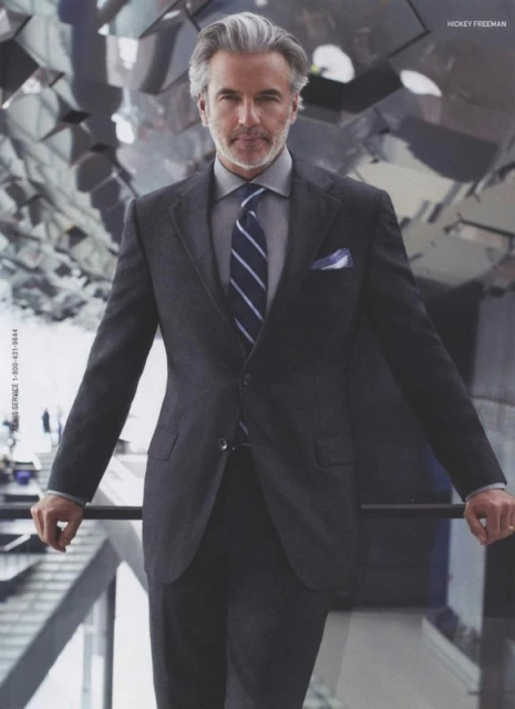 Portrait of a mature man in a dark grey suit, light grey shirt, and blue tie. He stands in an open, bright room.