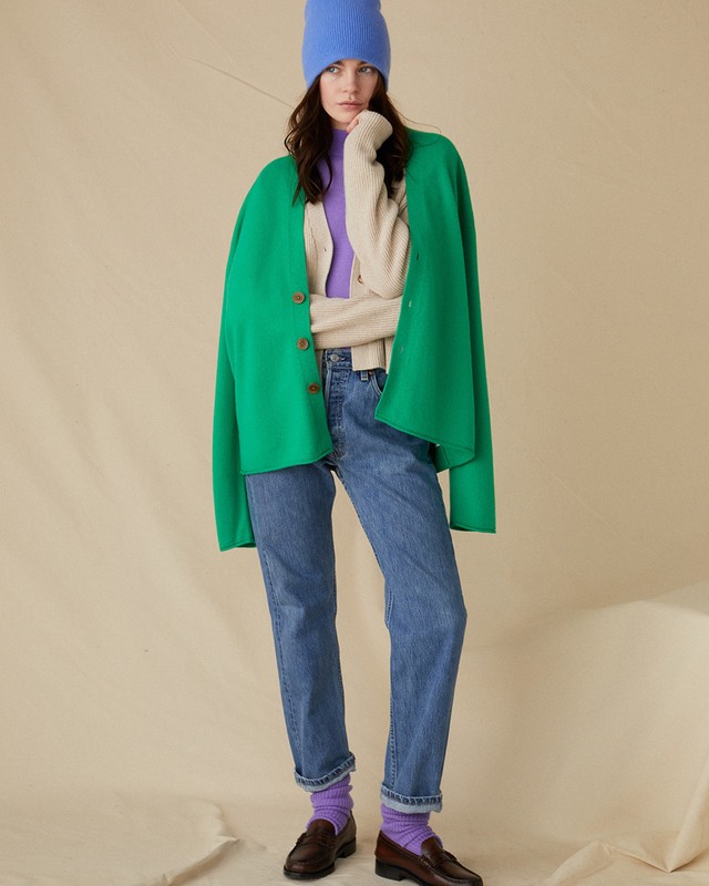 Fashion model posing in a studio wearing jeans, sweater, green coat, blue beanie, and brown loafers on a beige background.