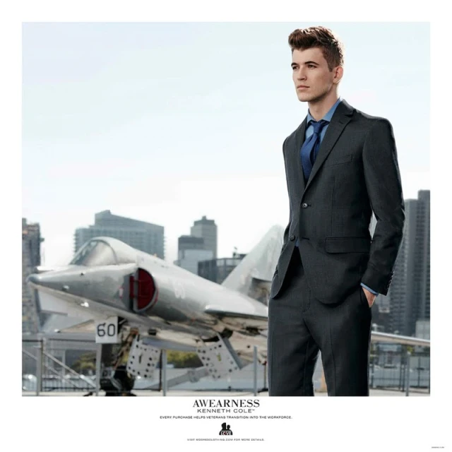 Man in suit stands near jet on aircraft carrier with city skyline backdrop, professional and polished look with a modern military vibe.
