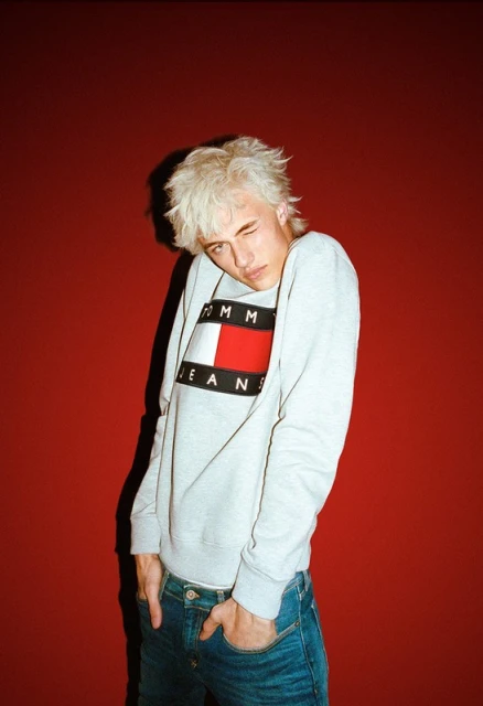 A young adult male with bleached hair and a Tommy Jeans sweatshirt poses against a red backdrop, winking at the camera in a casual style.