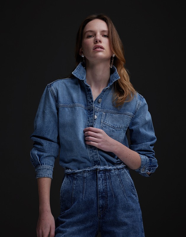 Fashionable adult woman with fair skin poses in a blue denim collared long-sleeved shirt and matching pants against a dark backdrop.