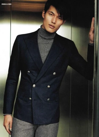 Male fashion model poses indoors wearing a dark blue double-breasted blazer, grey turtleneck, and grey trousers in an elevator setting.