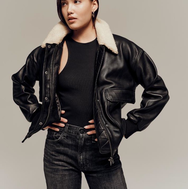 A woman poses in a black leather jacket with a light fur collar, black top, and black jeans against a neutral studio backdrop.