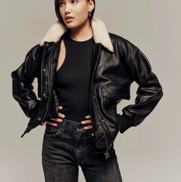 A woman poses in a black leather jacket with a light fur collar, black top, and black jeans against a neutral studio backdrop.