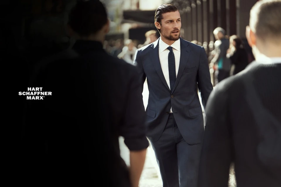 A handsome man in a blue suit walks confidently through a bustling urban setting, framed by blurred figures and natural light.