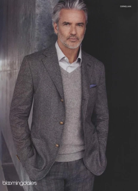 Mature man in grey suit jacket and sweater, posing indoors against a textured background, fashionable and sophisticated look.