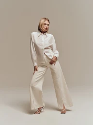 Blonde female model wearing a white button-down shirt and cream wide-leg trousers against a neutral studio background