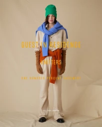 Model wearing cream cashmere jumpsuit, green beanie, blue sweater tied around shoulders, and orange sweater tied at waist against a textured beige backdrop