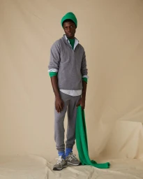 Black man in grey sweatshirt and sweatpants with green beanie holding green fabric against a textured seamless backdrop