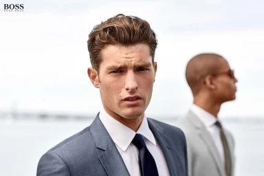 Close-up of a white man in a grey suit and dark tie with another man in the blurred background near water.