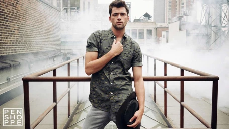 Man wearing a patterned olive short-sleeve shirt and grey pants holding a hat on a rooftop setting in Los Angeles.