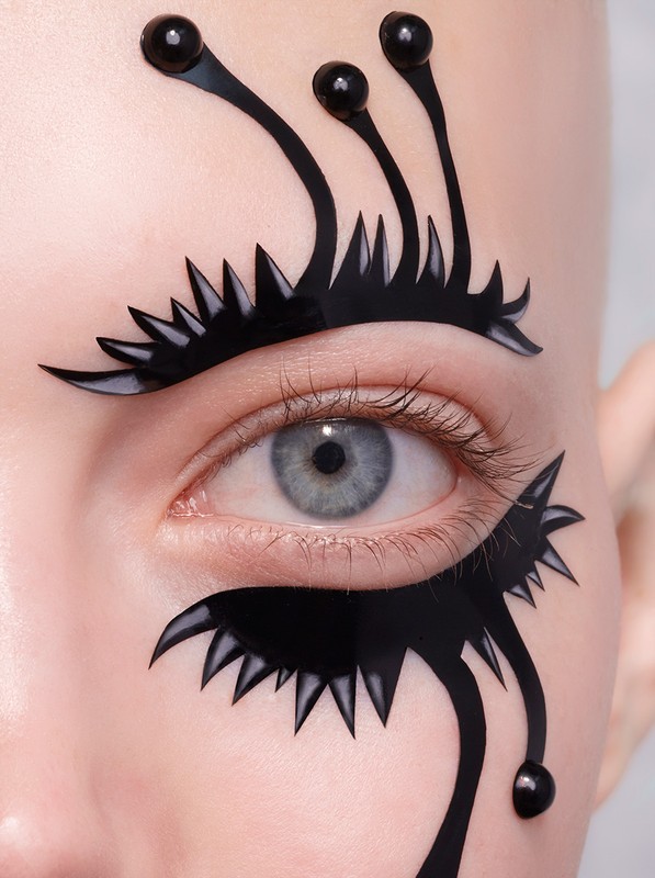 Close-up of a woman's face featuring decorative black adhesive designs around the eye, creating a stylized, artistic effect with gray eye.