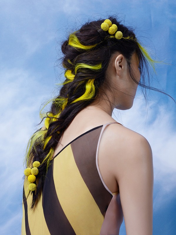 Brunette woman with yellow dyed hair in a braid, adorned with yellow floral accents against a blue sky background, in fashion shot.