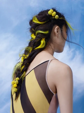 Brunette woman with yellow dyed hair in a braid, adorned with yellow floral accents against a blue sky background, in fashion shot.