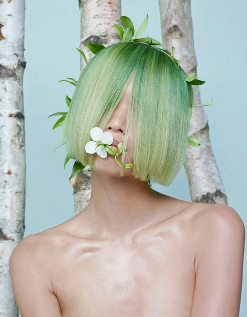 Young woman with creative green hair and leafy accents, holding a white flower in her mouth, posing against birch tree trunks.