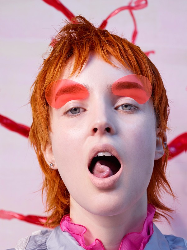 Close-up portrait of a young adult female with red hair and translucent red eyebrow covers against a light pink and white background.