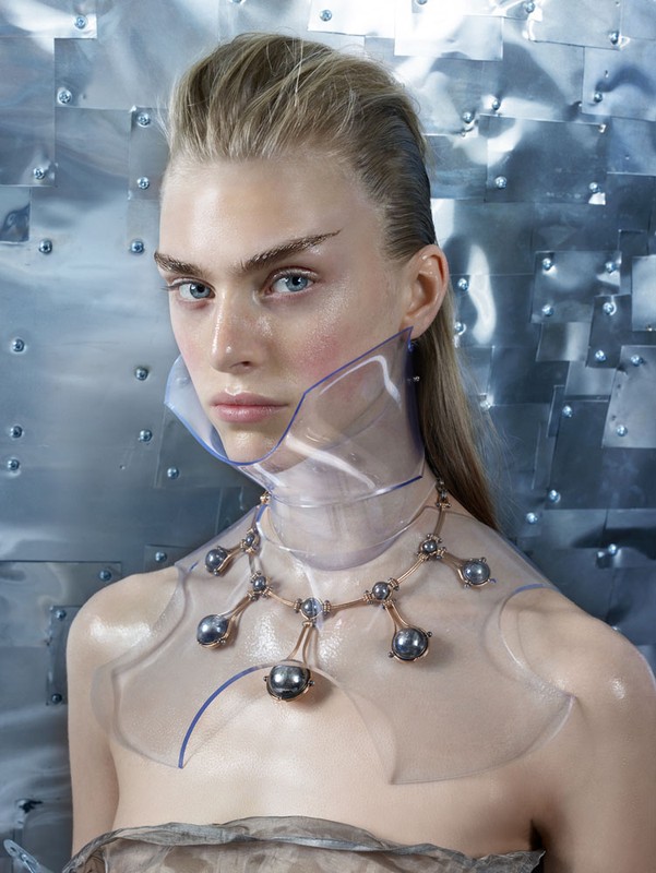 Striking beauty shot of a fair-skinned woman with light hair, wearing a futuristic clear plastic collar and ornate black pearl necklace.
