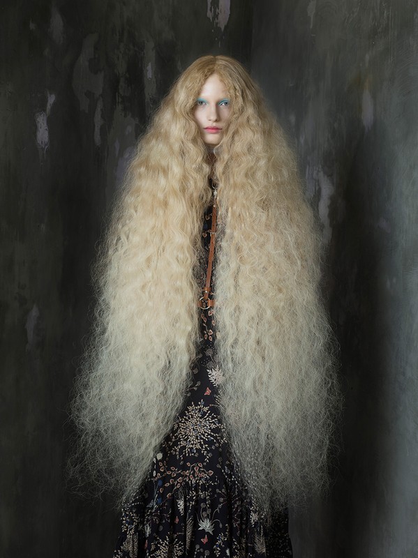 Indoor portrait of a female fashion model with an elaborate long blond wig, wearing a floral dress, set against a grungy grey background.