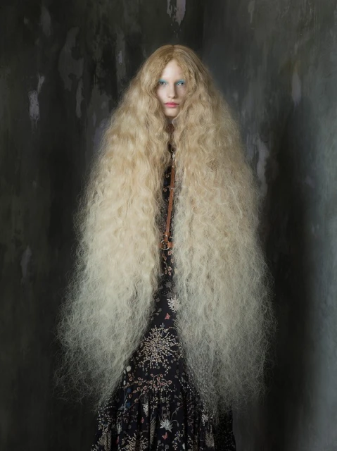 Indoor portrait of a female fashion model with an elaborate long blond wig, wearing a floral dress, set against a grungy grey background.