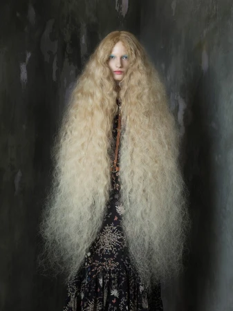 Indoor portrait of a female fashion model with an elaborate long blond wig, wearing a floral dress, set against a grungy grey background.