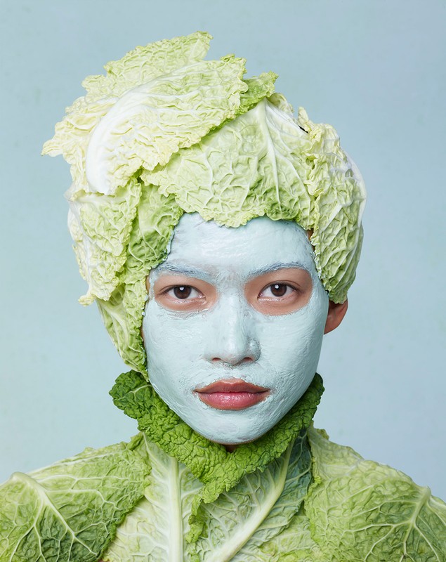 Close-up of a person with light skin and a light-green face mask, wearing a head covering and garment made of savoy cabbage leaves.