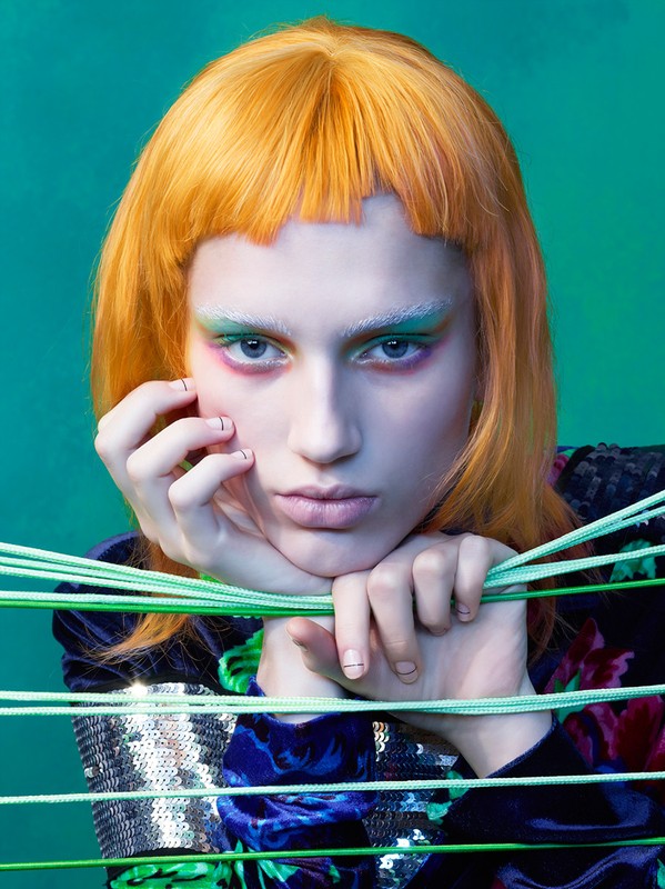 Close-up portrait of a young adult female with orange hair and bright, colorful makeup, posing with horizontal green string lines.