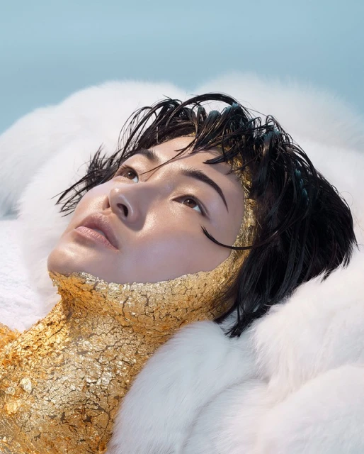 A woman with short black hair and golden metallic face and body paint lies against fluffy white faux fur in a beauty shot against a light blue backdrop.