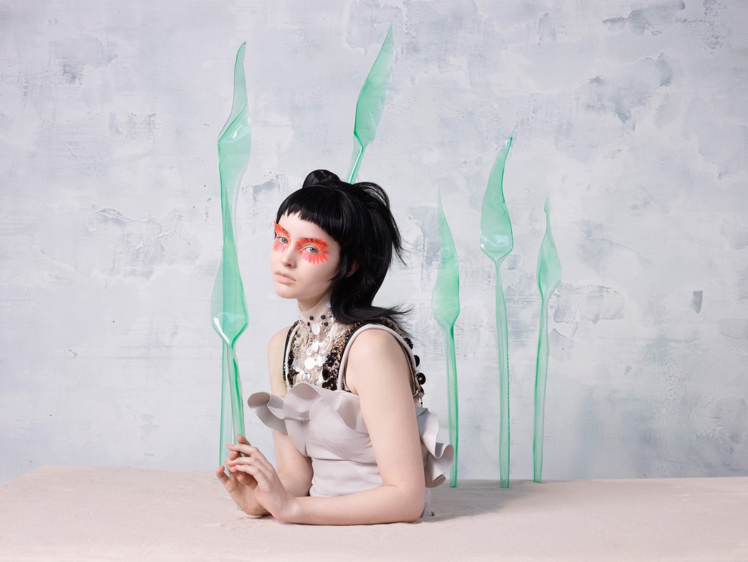 Woman with artistic eye makeup poses at a light peach table surrounded by translucent green plastic utensils in a studio.