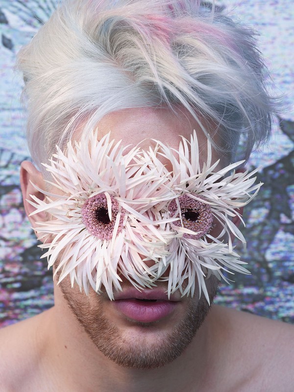 Close-up of a person with white hair and a light beard, partially obscured by white daisy-like flowers covering the eyes.