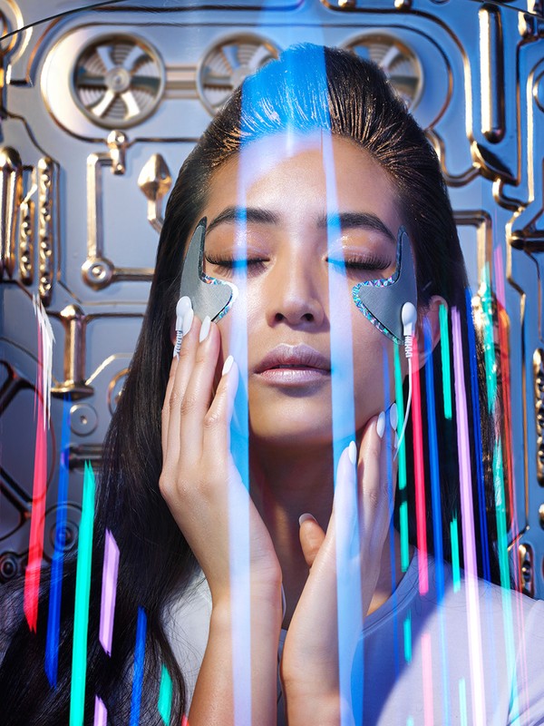 Close-up of a woman with closed eyes, wearing silver eye patches, and standing in front of a futuristic metallic background.