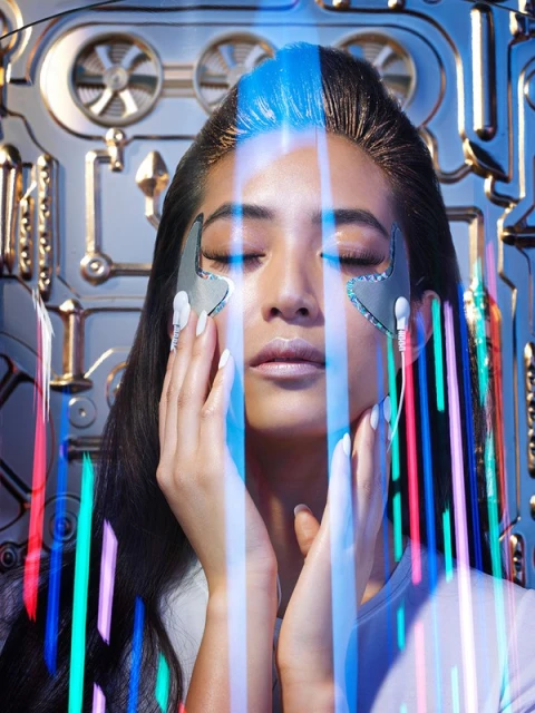 Close-up of a woman with closed eyes, wearing silver eye patches, and standing in front of a futuristic metallic background.