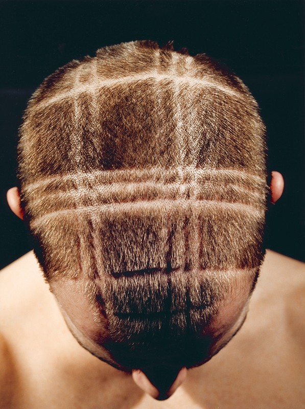 Close-up of the back of a person's head with short brown hair, featuring a unique grid pattern shaved into the hair, against a dark backdrop.
