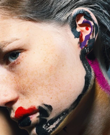 Close-up portrait of a woman with freckles and painted colorful abstract marks on her face and ear by Michael Baumgarten