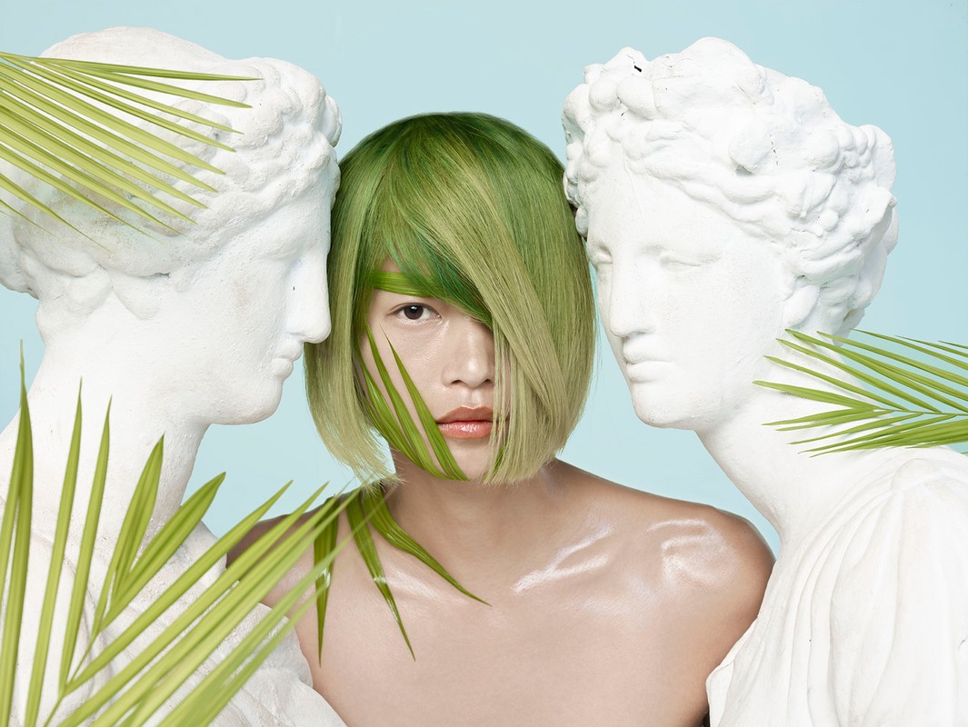 Artistic portrait of a woman with green hair standing between two statues with palm fronds against a pale blue backdrop in a studio.
