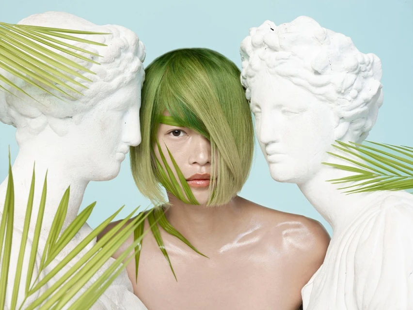 Artistic portrait of a woman with green hair standing between two statues with palm fronds against a pale blue backdrop in a studio.