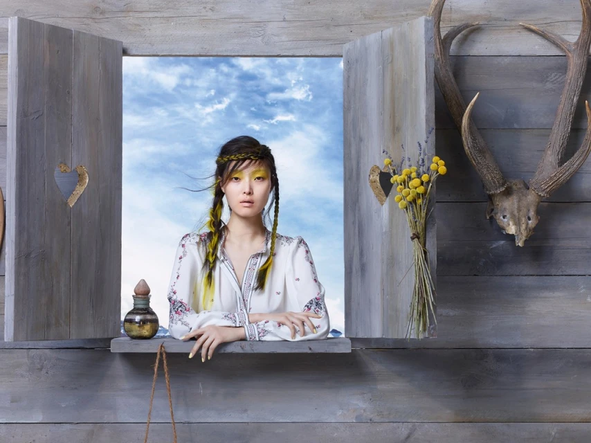 Young woman with braids and white embroidered blouse frames blue skies as she poses inside a rustic window beside a deer skull.