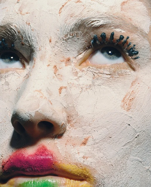 Close-up of a person's face covered in cracked white clay, dramatic blue mascara, and vibrant rainbow lipstick, creating an abstract beauty look.
