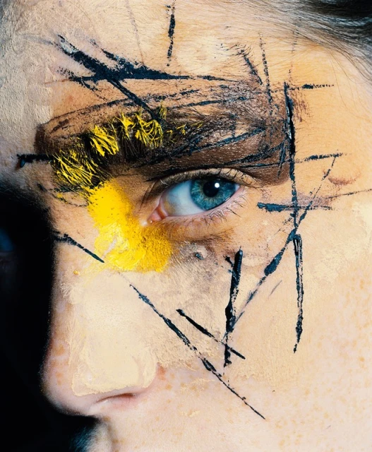 Portrait of a person with striking face paint. Yellow and black paint accents the face and eye, creating an artistic and bold visual statement.