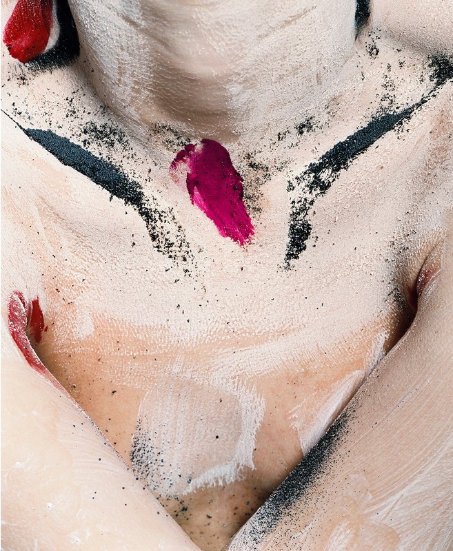 Close-up shot of a model's torso covered in white paint and black powder, accented with vivid red paint streaks, creating a textured, artistic effect.