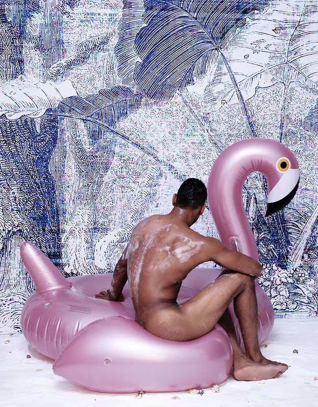 Nude man sits on a pink flamingo inflatable, tropical foliage background. Body is slightly turned, gazing away in a studio setting.